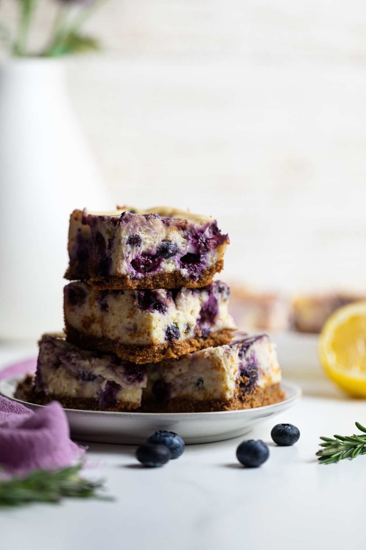 Lemon Blueberry Cheesecake Bars in a stack on a plate with fresh rosemary.