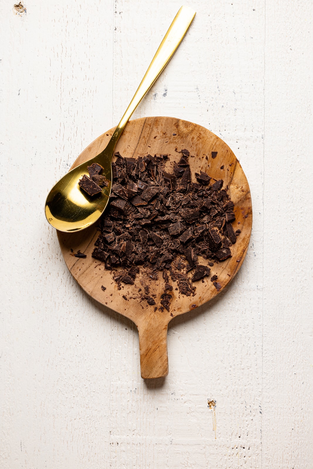 Chopped chocolate on a wood plate on a white wood table with a gold spoon.