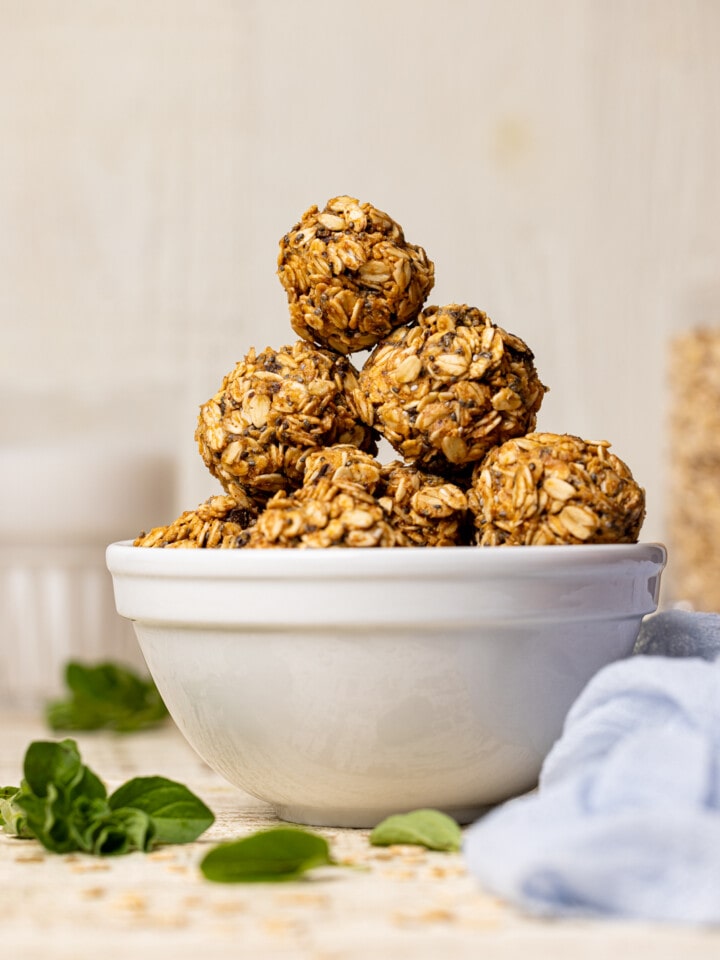 Protein balls stacked on each other in a white bowl on a white wood table with herbs and a light blue napkin.