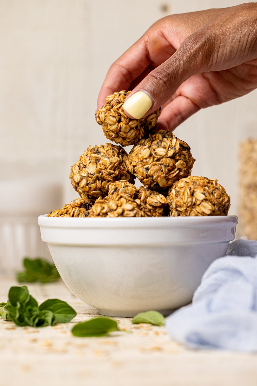 Protein balls stacked on each other in a white bowl on a white wood table with herbs and a light blue napkin and one ball being picked up.