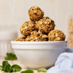 Protein balls stacked on each other in a white bowl on a white wood table with herbs and a light blue napkin.