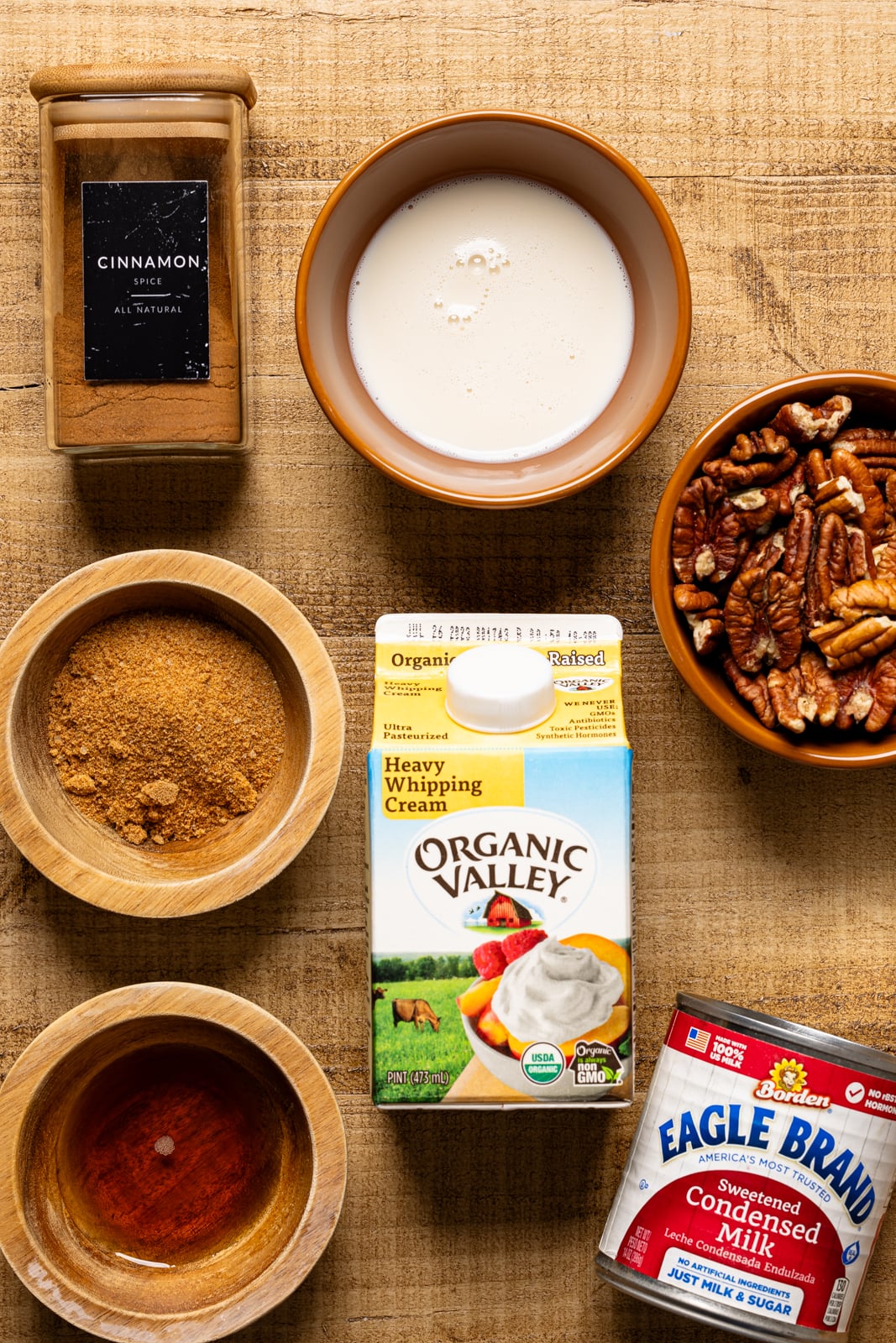 Ingredients on a brown wood table including heavy cream, cinnamon, sugar, condensed milk, pecans, and maple syrup.