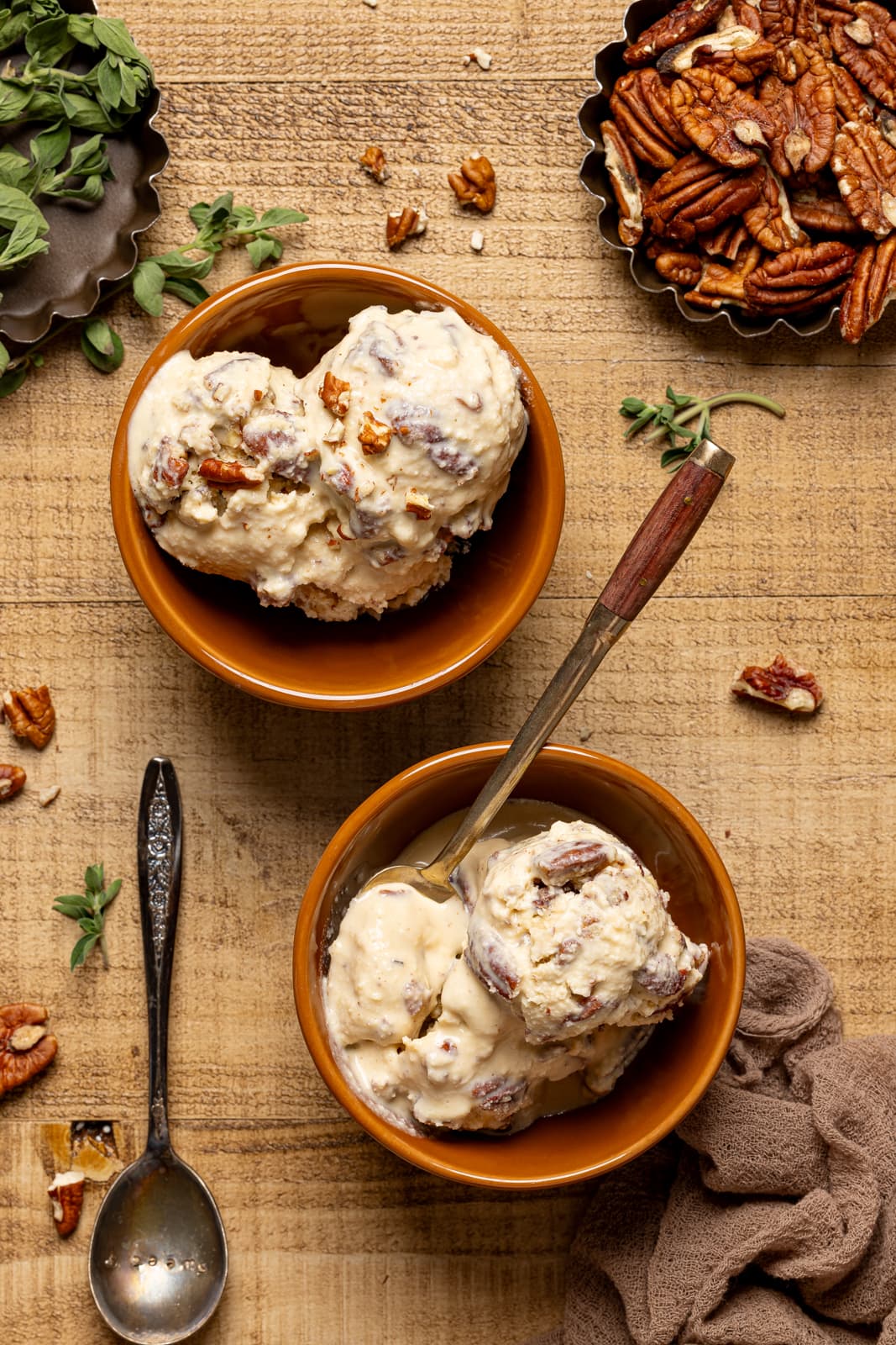Two bowls of ice cream on a brown wood table with spoons and chopped pecans.