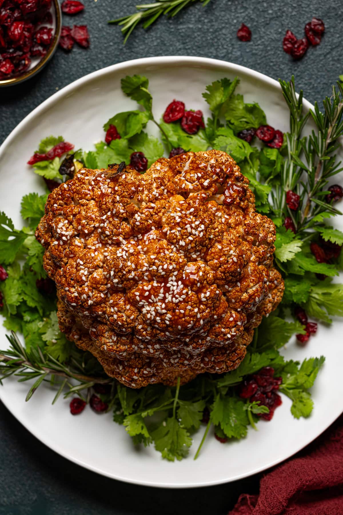 Whole roasted cauliflower in a white serving platter with garnish of herbs + cranberries. 