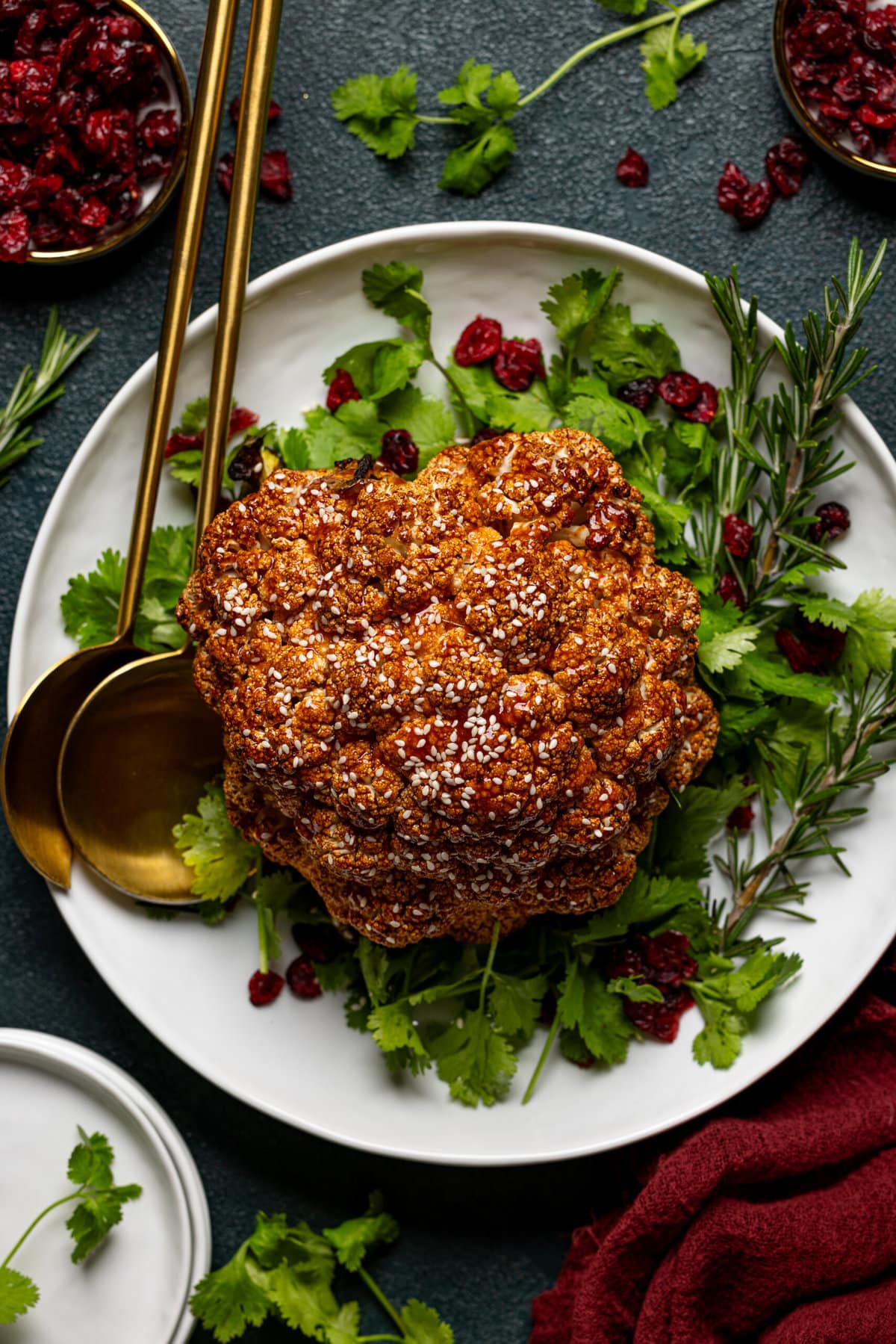 Whole roasted cauliflower atop herbs and a garnish with cranberries and two serving spoons. 