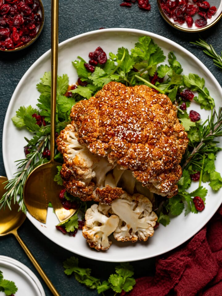Roasted cauliflower sliced atop herbs + cranberries on a platter with two serving spoons.