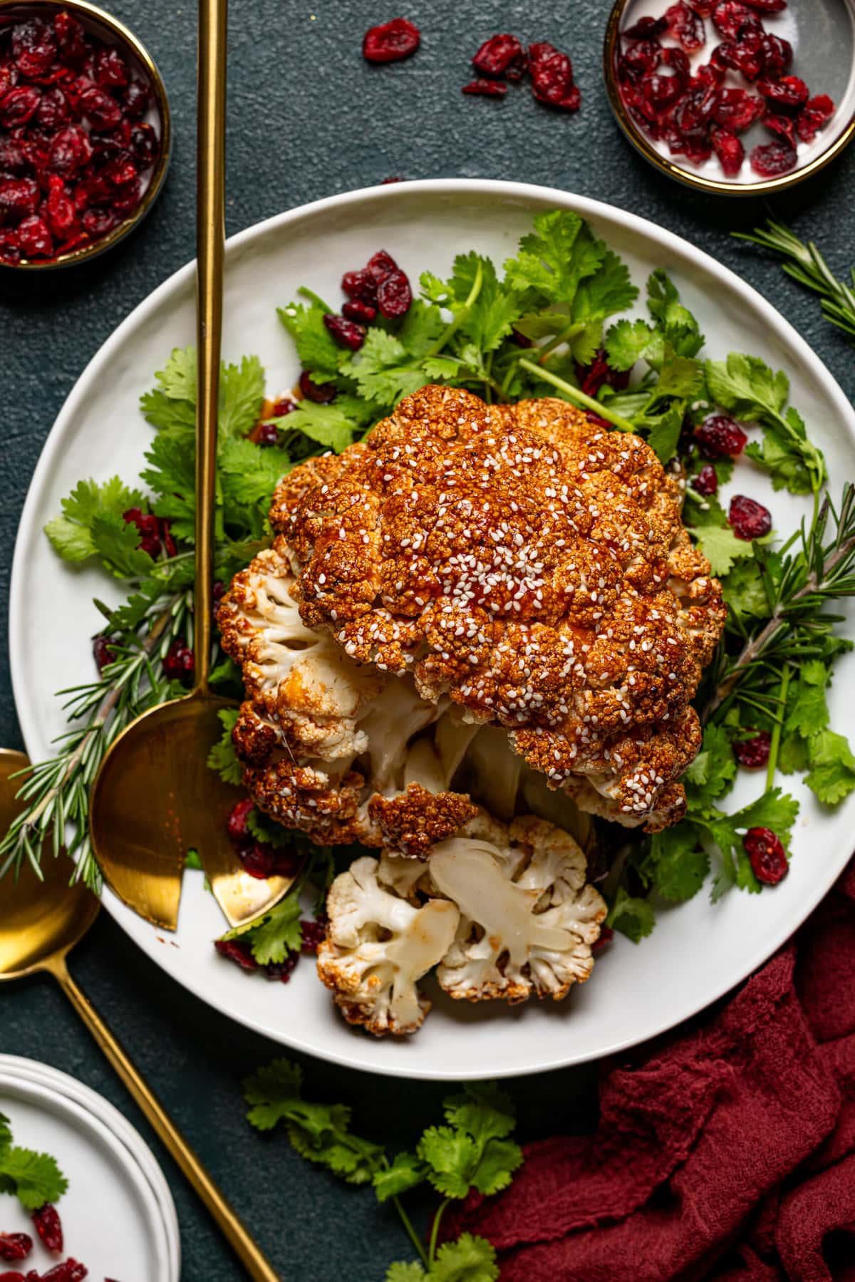 Roasted cauliflower sliced atop herbs + cranberries on a platter with two serving spoons.
