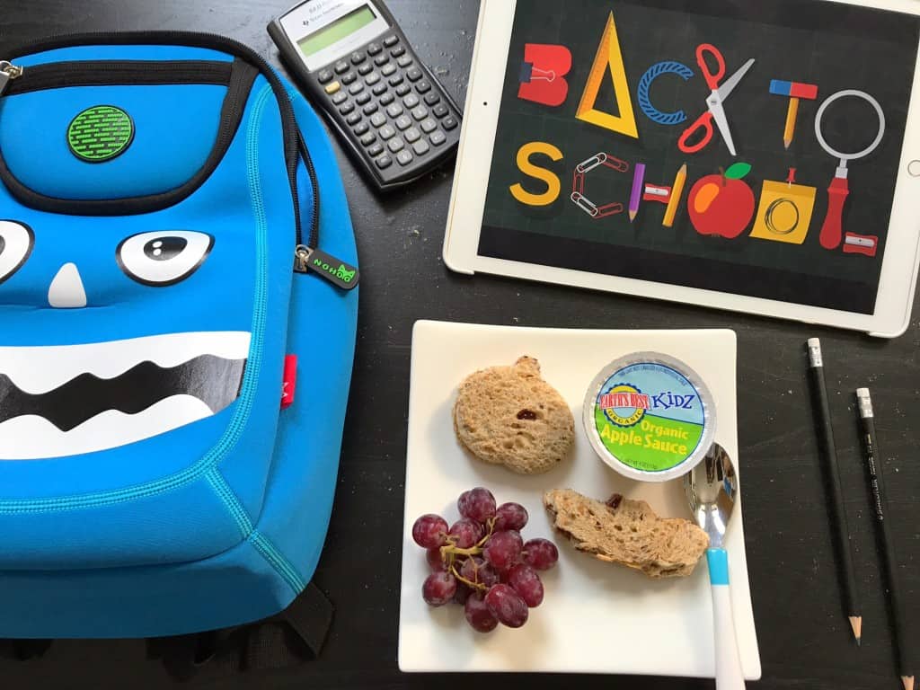 Blue lunch box next to a calculator, back to school sign, and food including grapes and apple sauce.