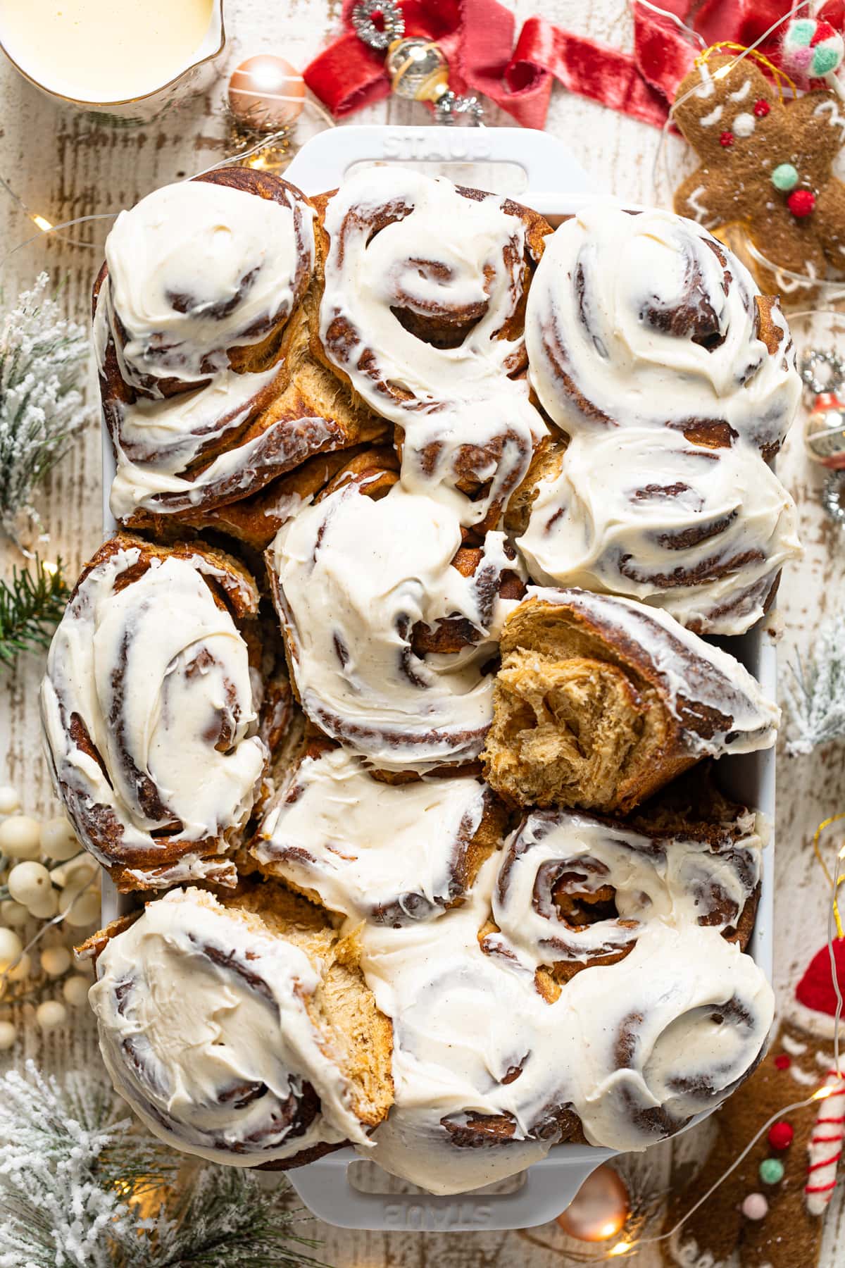 Gingerbread Cinnamon Rolls with Maple Cream Cheese Frosting in a baking pan surrounded by Christmas decorations