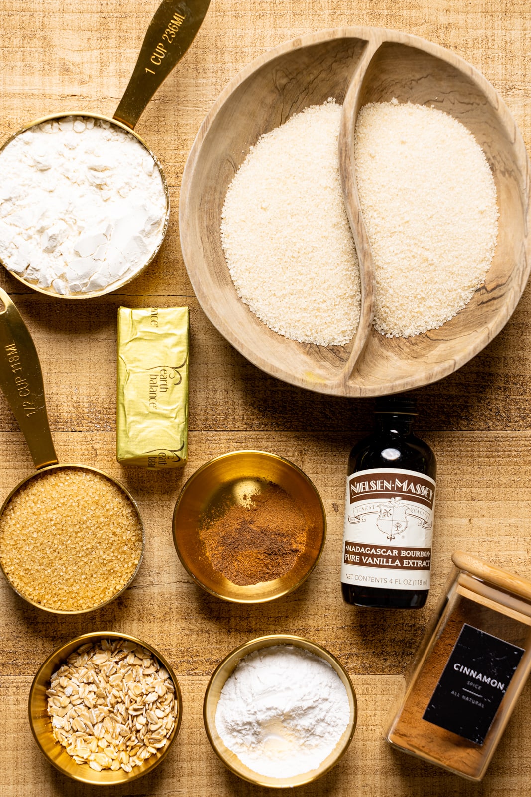 Ingredients on a brown wood table including sugar, flour, sugar, spices, oats, and vanilla.