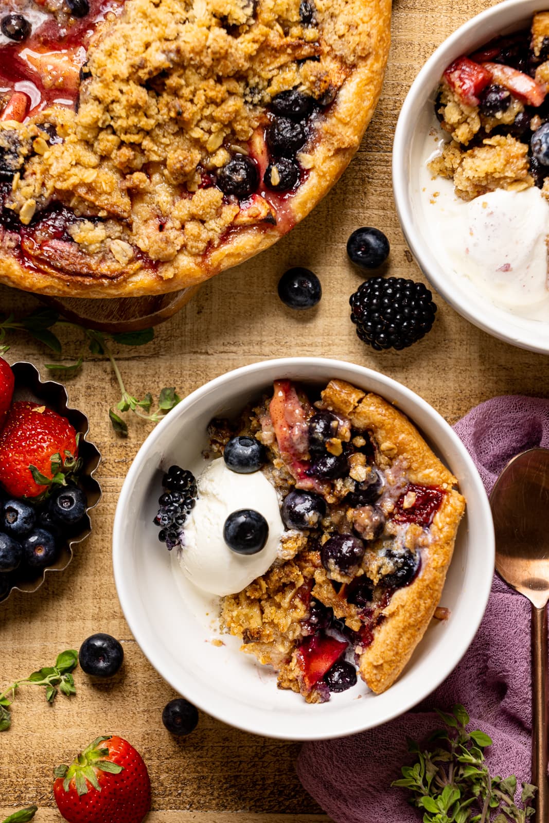 Pie slice in a white bowl on a brown wood table with a scoop of ice cream and berries alongside baked pie.