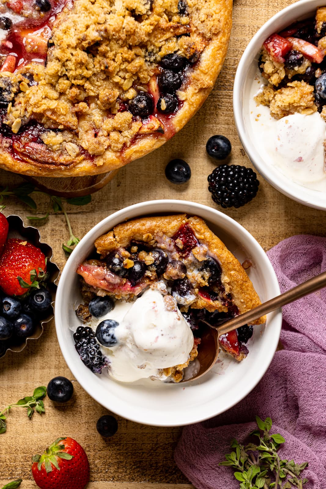Pie slice in a white bowl on a brown wood table with a scoop of ice cream and berries.