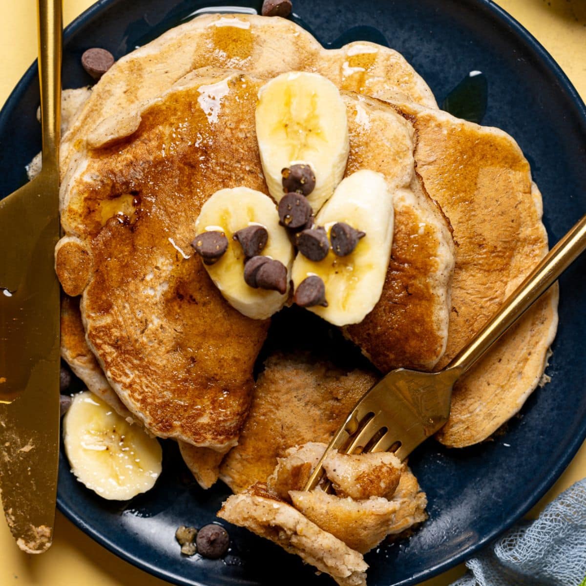 Up close shot of pancakes in a plate with fork + knife and topped with banana slices + chocolate chips.