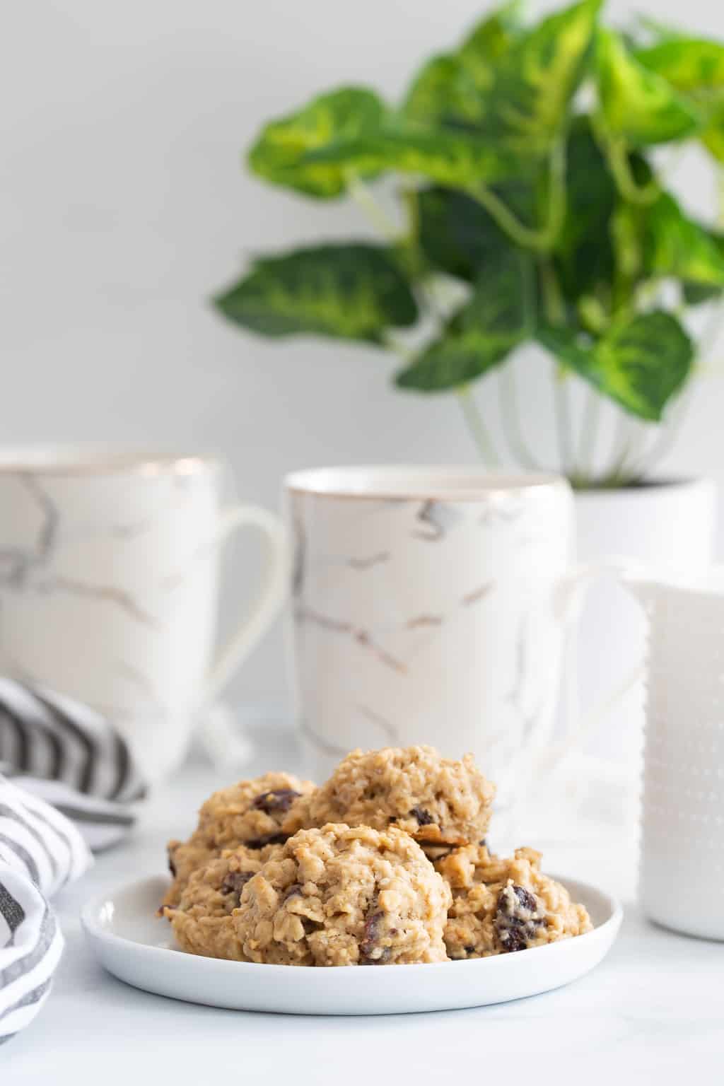 Plate of Chewy Apple Oatmeal Raisin Breakfast Cookies on a table with white mugs.