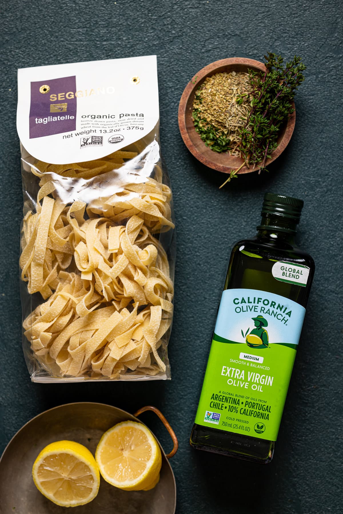 Ingredients on a dark green table including olive oil, pasta, herbs + seasonings, and lemon slices.