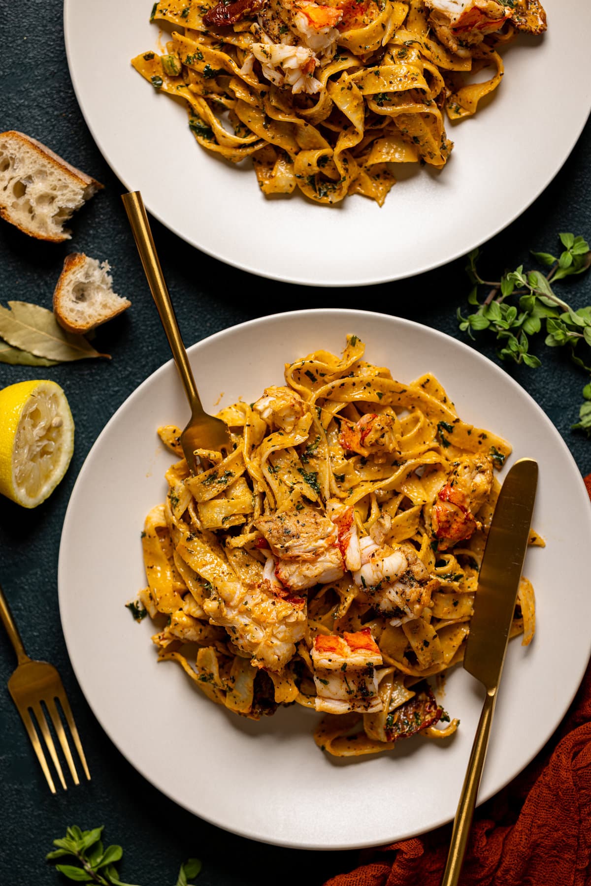 Tuscan lobster pasta in a plate with a fork and knife on a dark green table surrounded by garnish of lemons, bread, and herbs. 