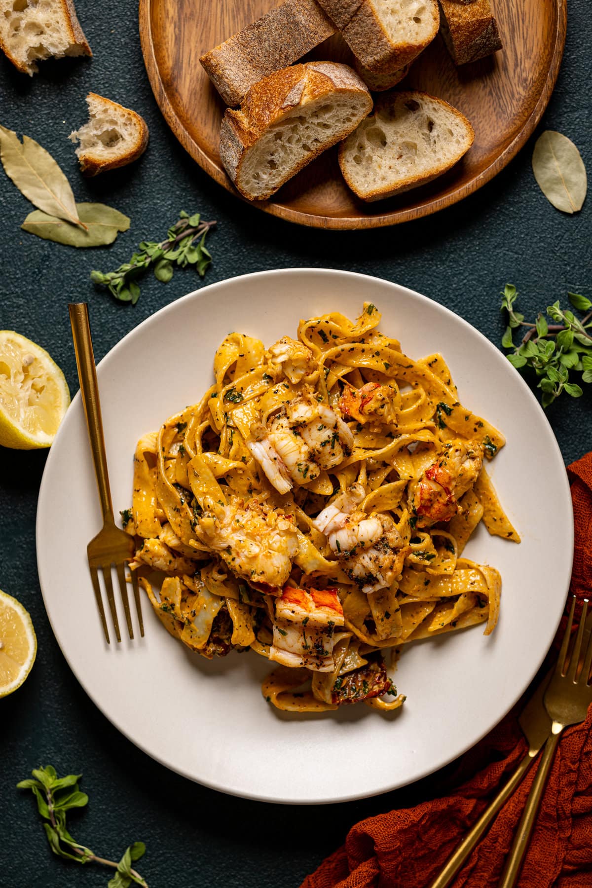 Tuscan lobster pasta in a plate with a fork on a dark green table with two gold forks and surrounded by garnish of lemons, plate of bread, and herbs. 