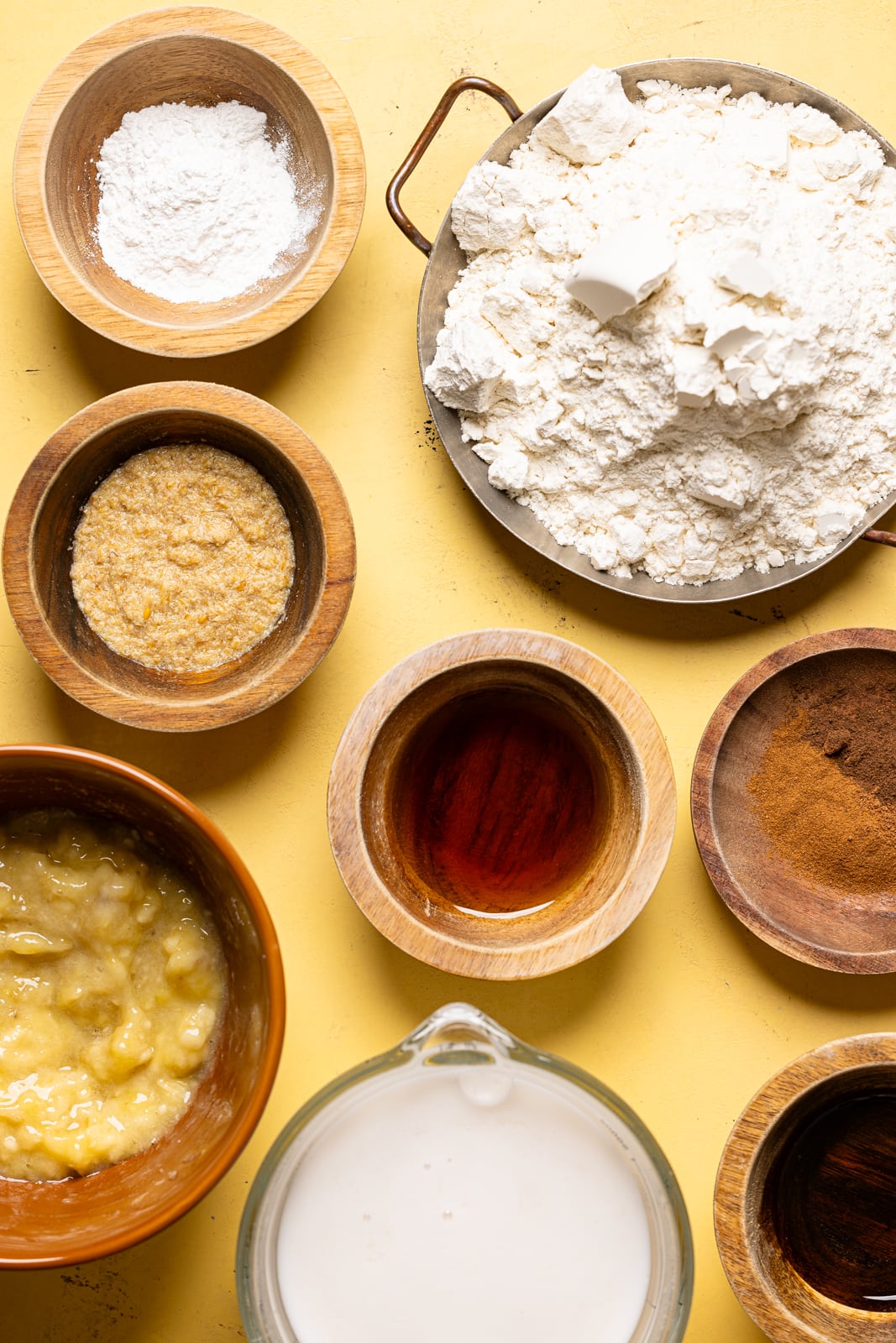 Ingredients on a yellow table including flour, baking powder, mashed bananas, milk, spices, and flax 'eggs'. 