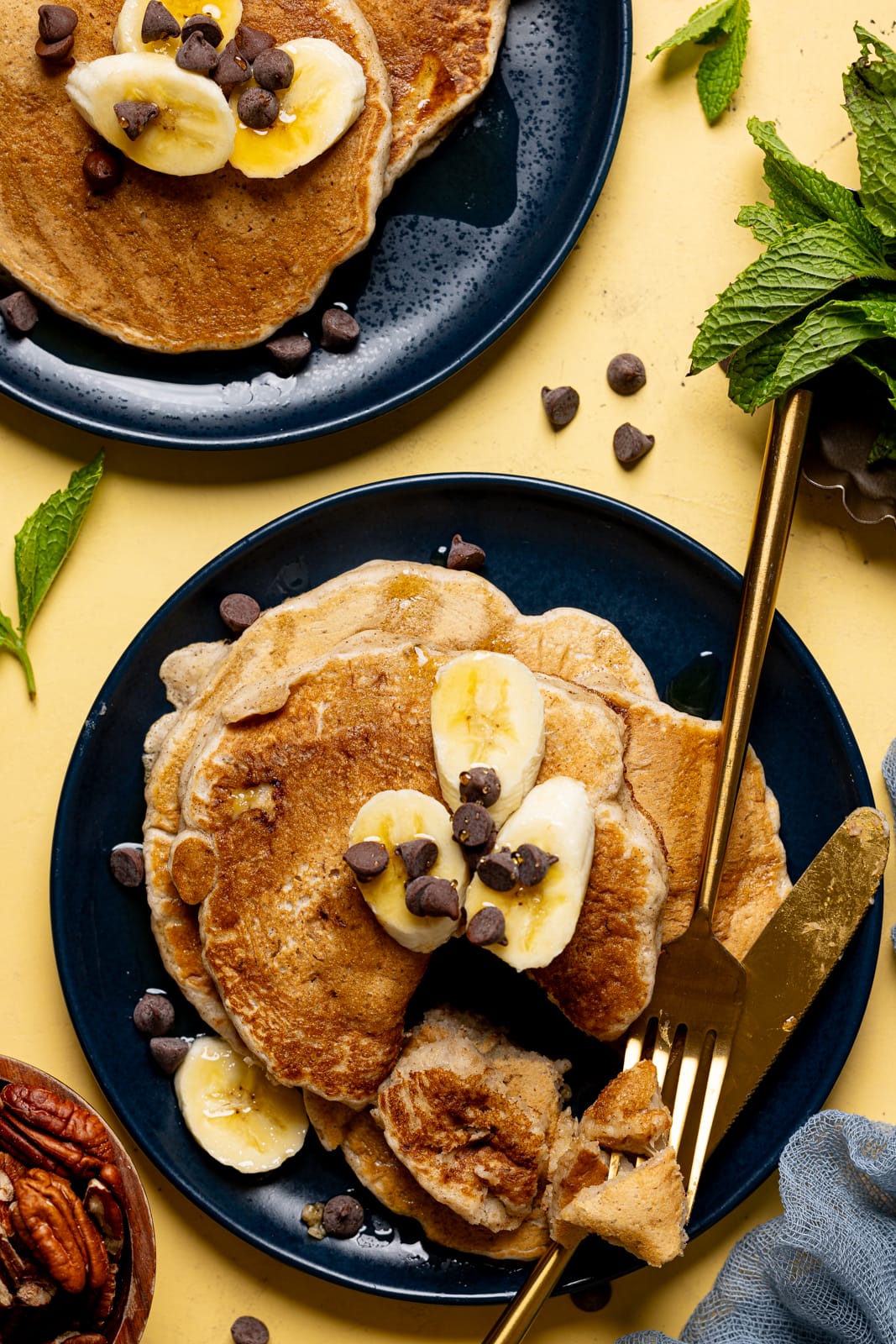 Pancakes on two plates with sliced bananas, chocolate chips, and sliced with a knife and fork.