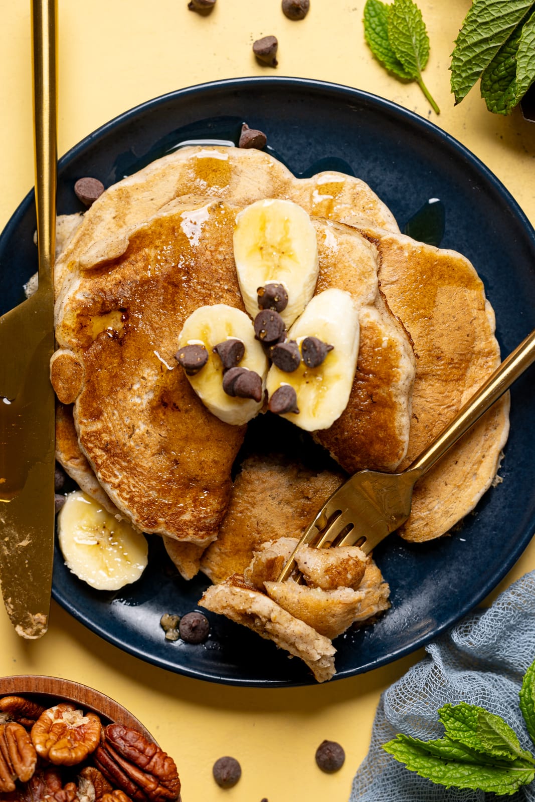 Up close shot of pancakes in a plate with fork + knife and topped with banana slices + chocolate chips.