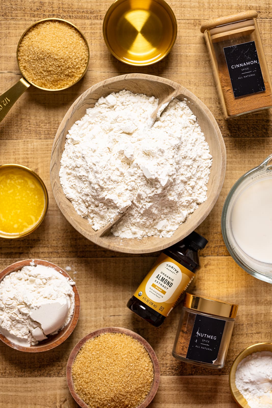 Ingredients on a brown wood table including flour, baking powder, sugar, spices, butter, almond extract, and milk.