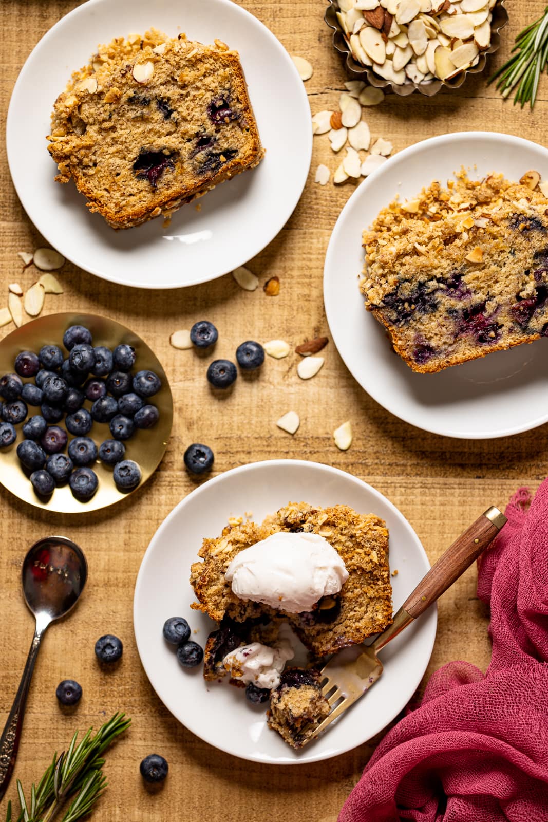 Banana bread on a brown wood table on three white plates with fresh blueberries, a spoon and fork, and a spoon of whipped cream.