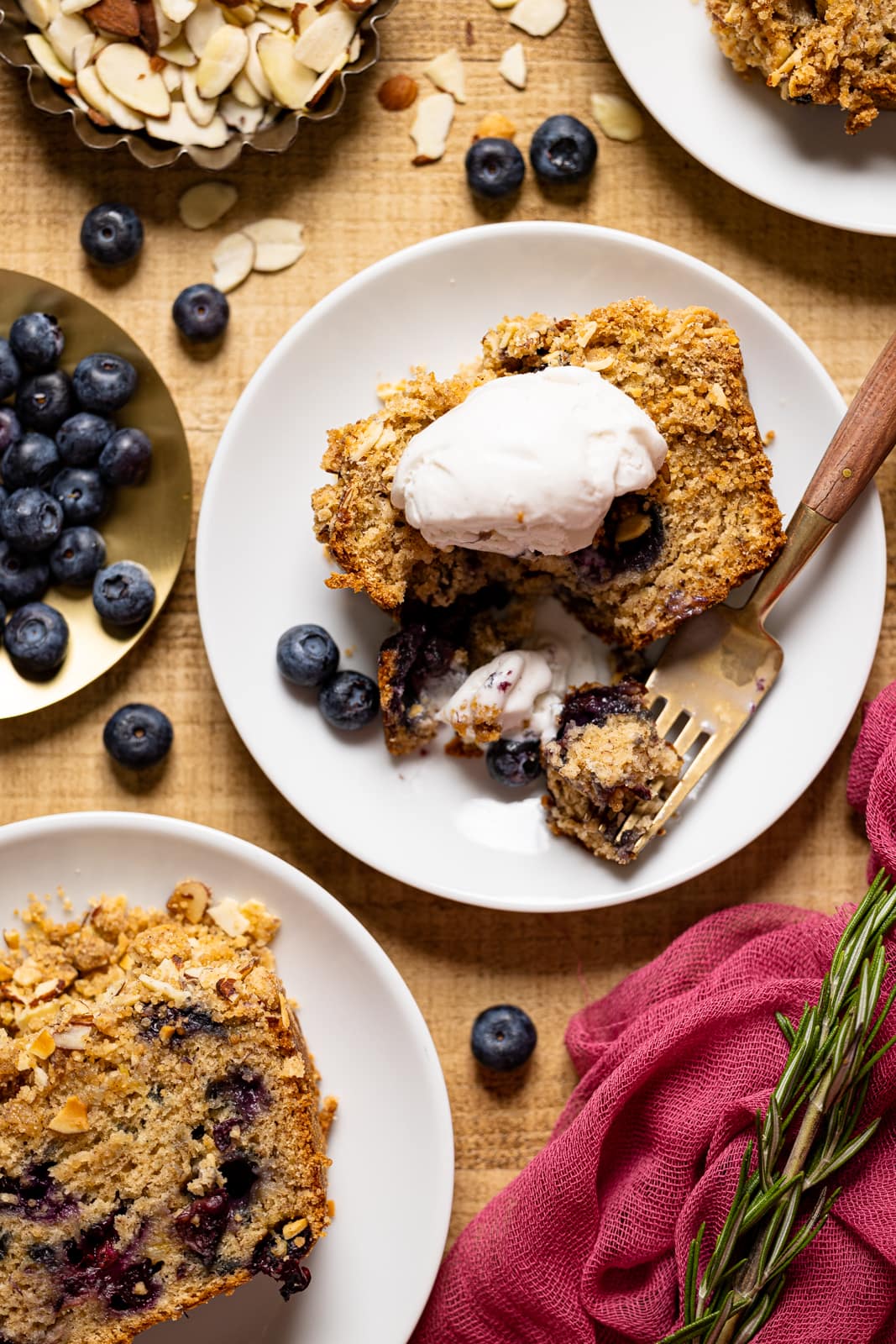 Banana bread on a brown wood table on three white plates with fresh blueberries and a spoon of whipped cream.