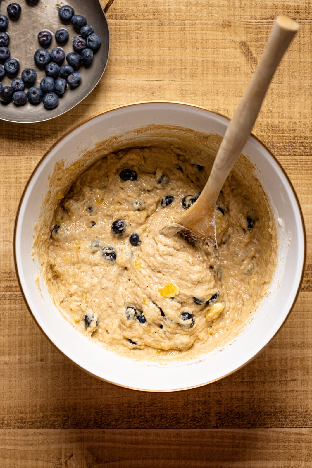 Bread batter in a large white bowl with blueberries and a wooden spoon on a brown wood table.
