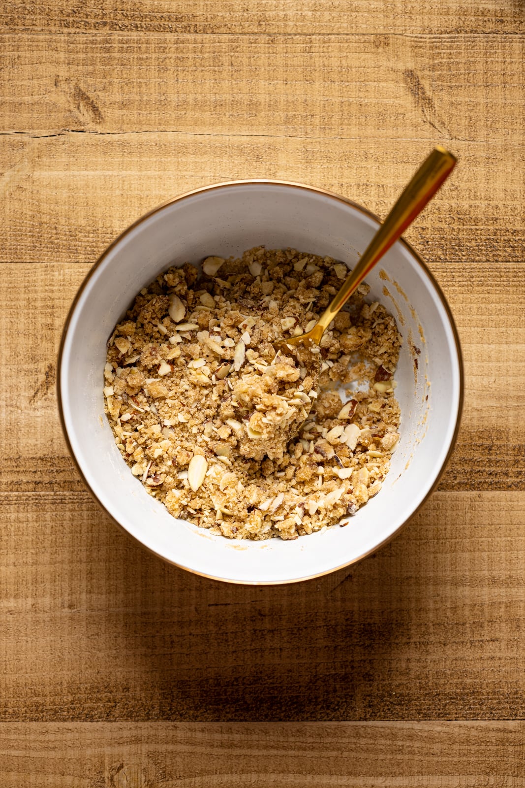 Crumble topping on a white bowl and a spoon on a brown wood table.