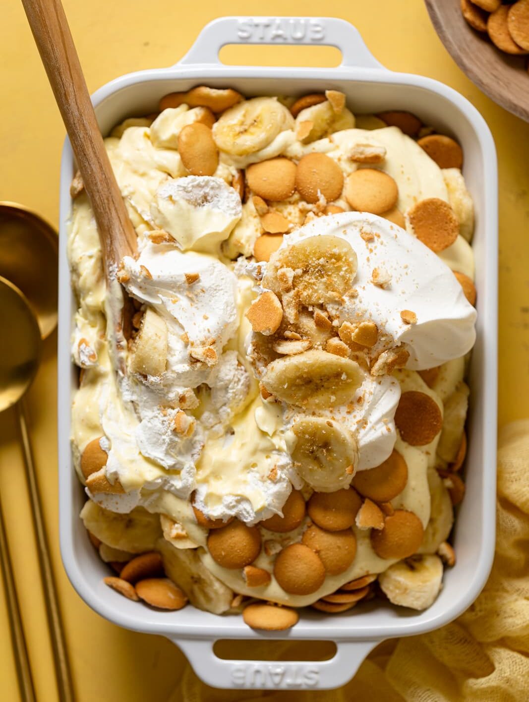 Banana Pudding in a white baking dish with a wooden spoon on a yellow table.