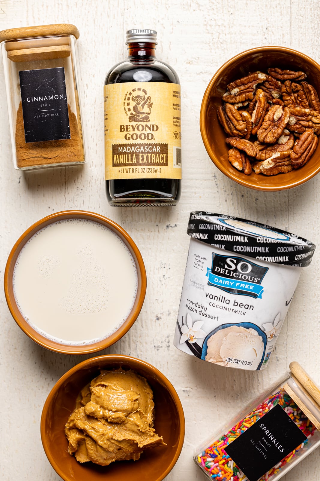 Ingredients on a white table including ice cream, milk, vanilla, cinnamon, pecans, peanut butter, and sprinkles.