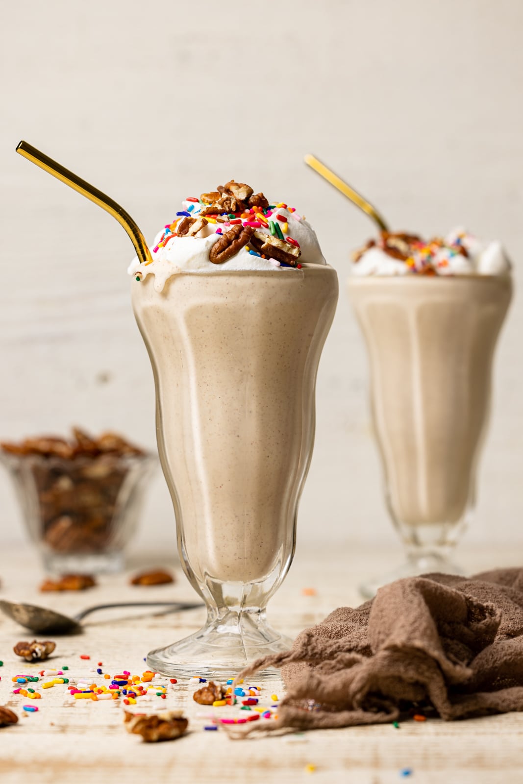 Two glasses of milkshake on a white table with a brown napkin, spoon, chopped pecans, and rainbow sprinkles.