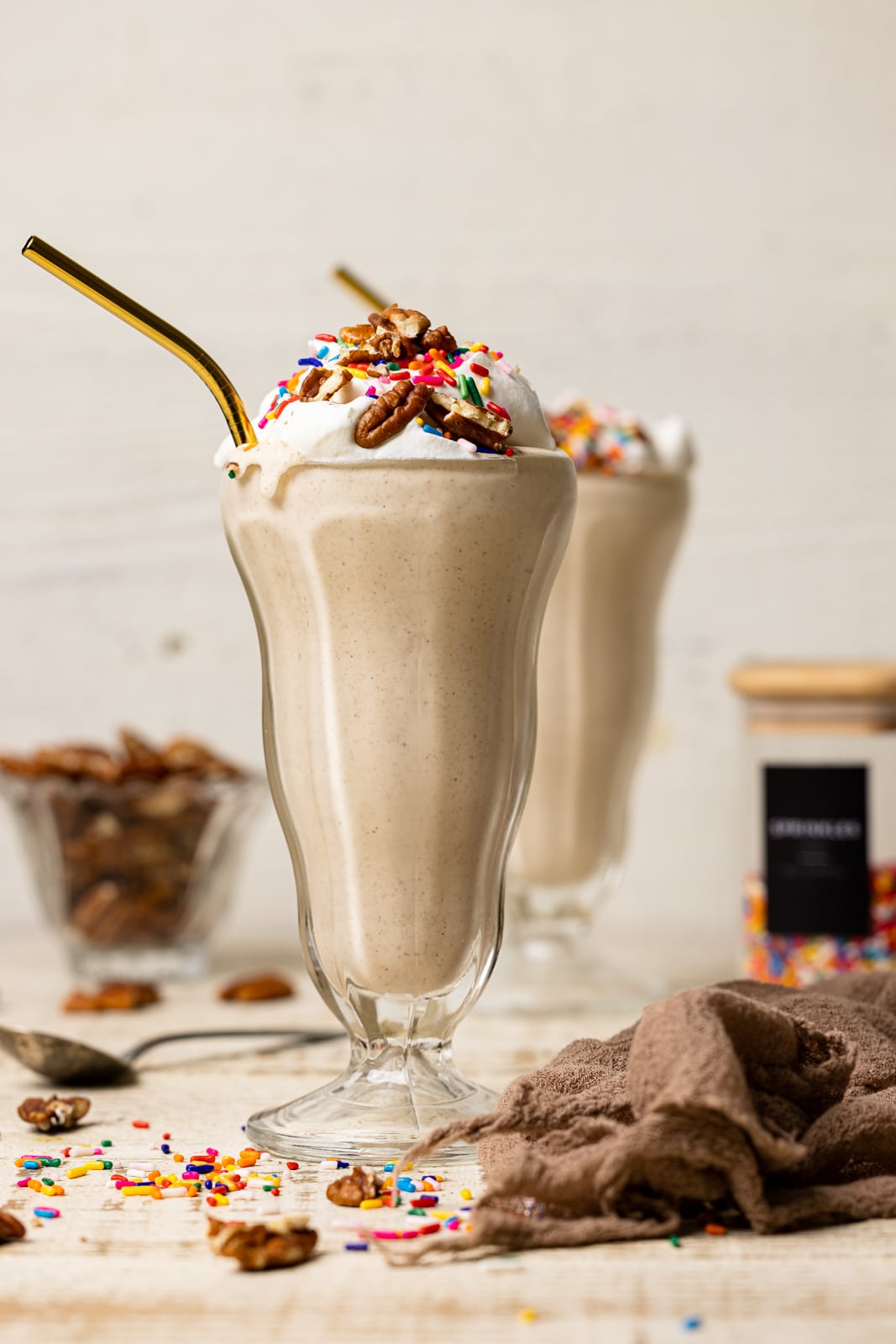 Two glasses of milkshake on a white table with a brown napkin, spoon, chopped pecans, and rainbow sprinkles.