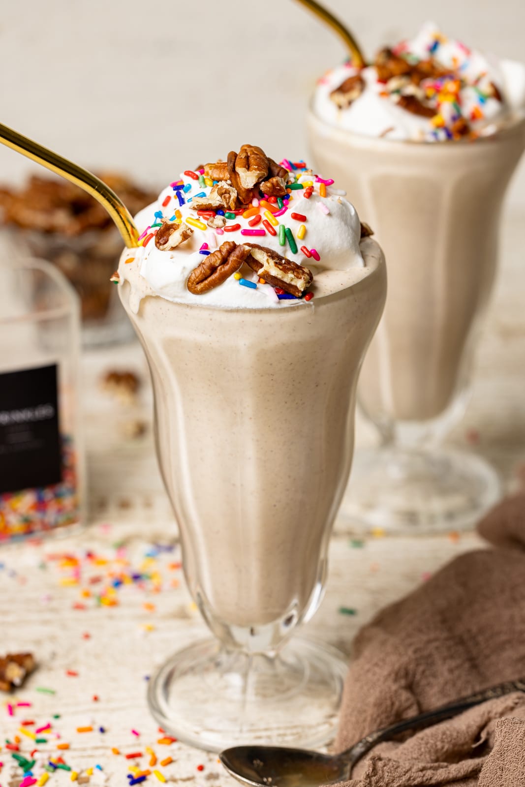 Two milkshakes in a glasses on a white table with rainbow sprinkles and pecans.