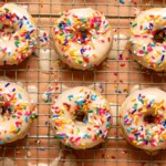 Up close shot of frosted donuts with rainbow sprinkles on a wire rack on a pink table.