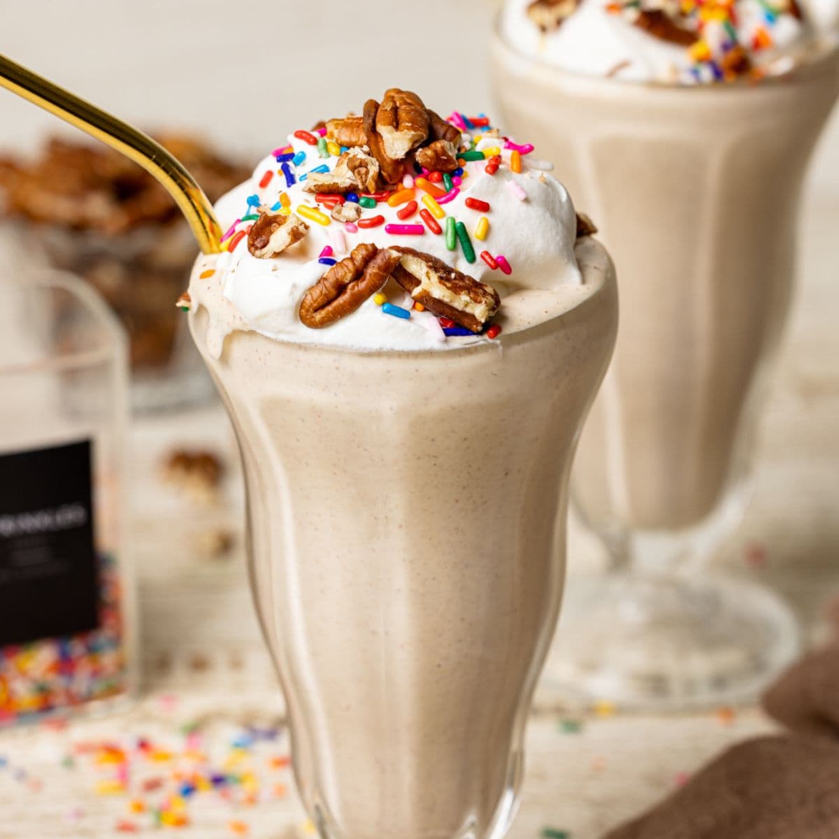 Two milkshakes in a glasses on a white table with rainbow sprinkles and pecans.