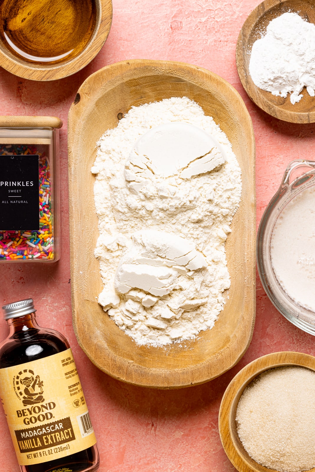 Ingredients on a pink table including flour, milk, sugar, rainbow sprinkles, vanilla, baking powder, and oil.