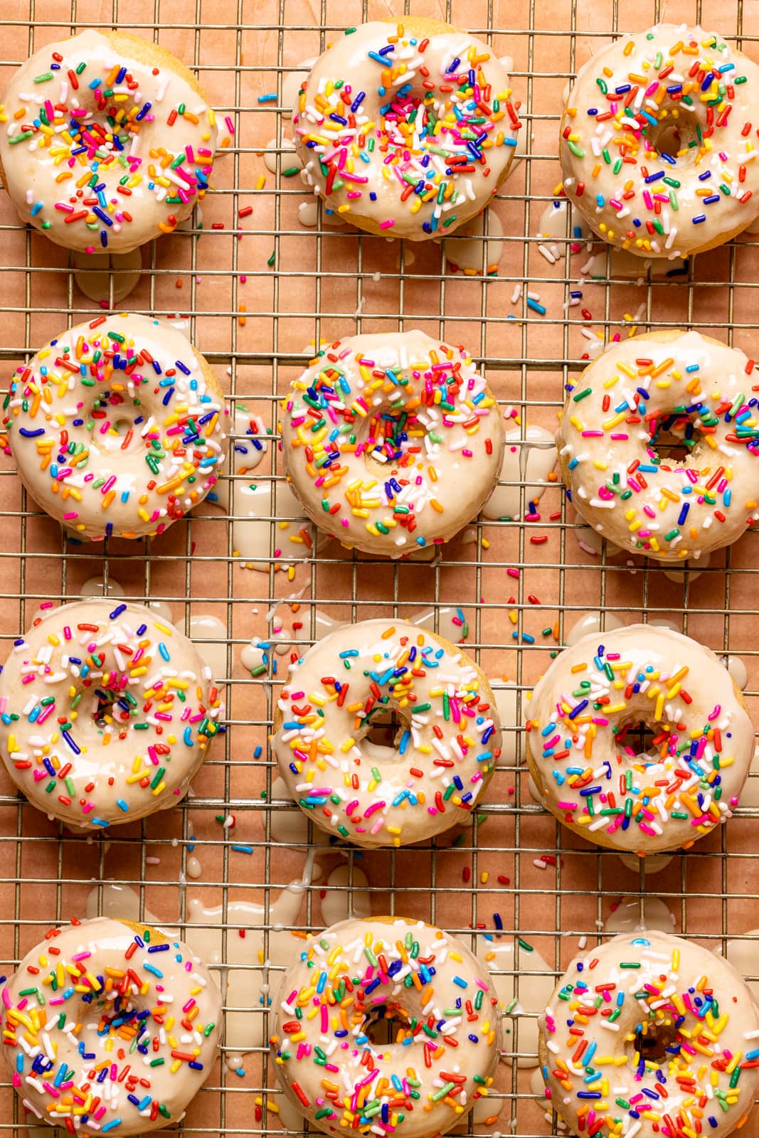 Frosted donuts with rainbow sprinkles on a wire rack on a pink table.