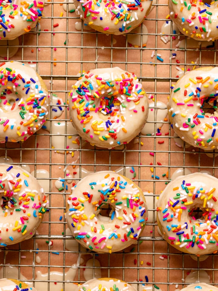 Up close shot of frosted donuts with rainbow sprinkles on a wire rack on a pink table.