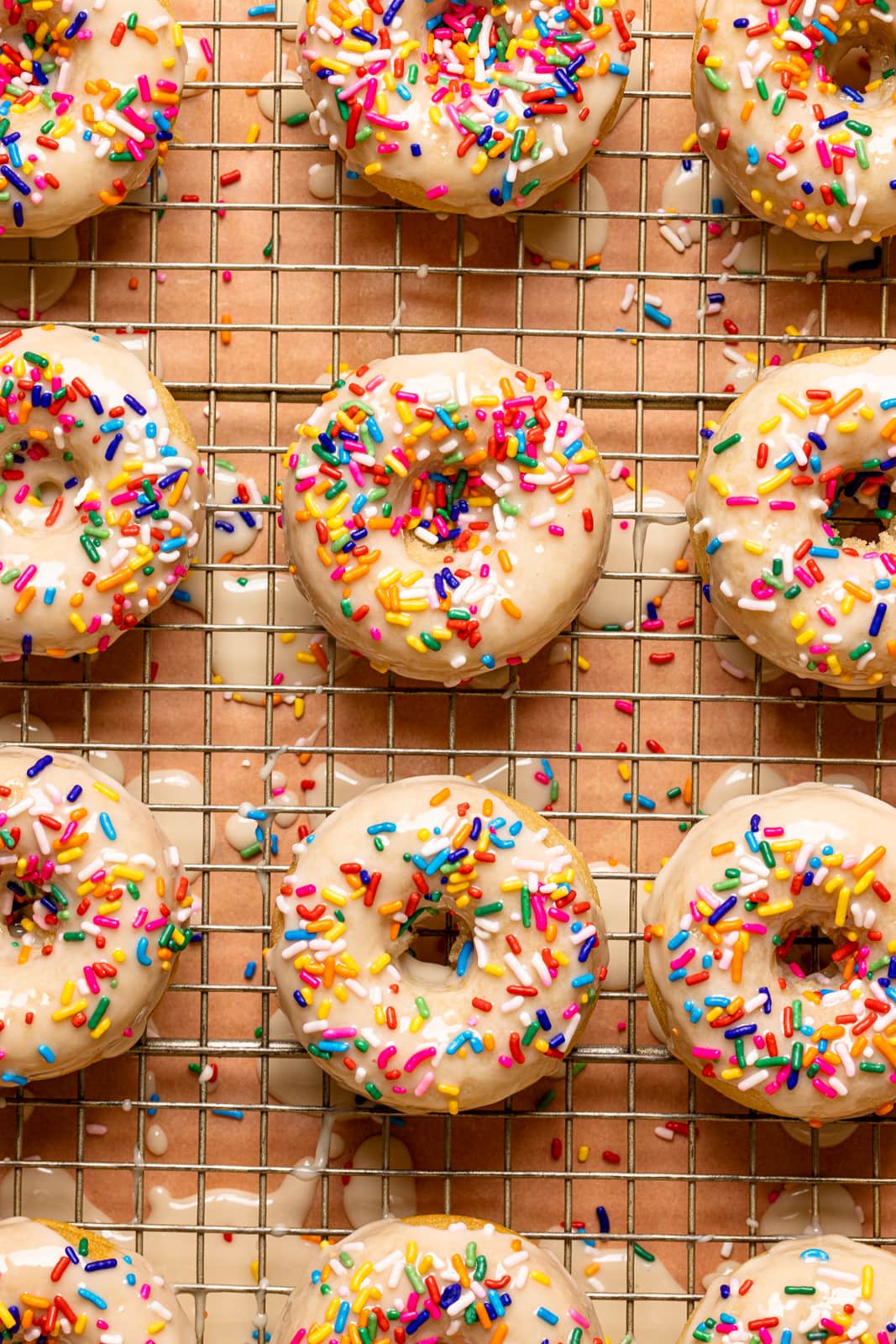 Up close shot of frosted donuts with rainbow sprinkles on a wire rack on a pink table.