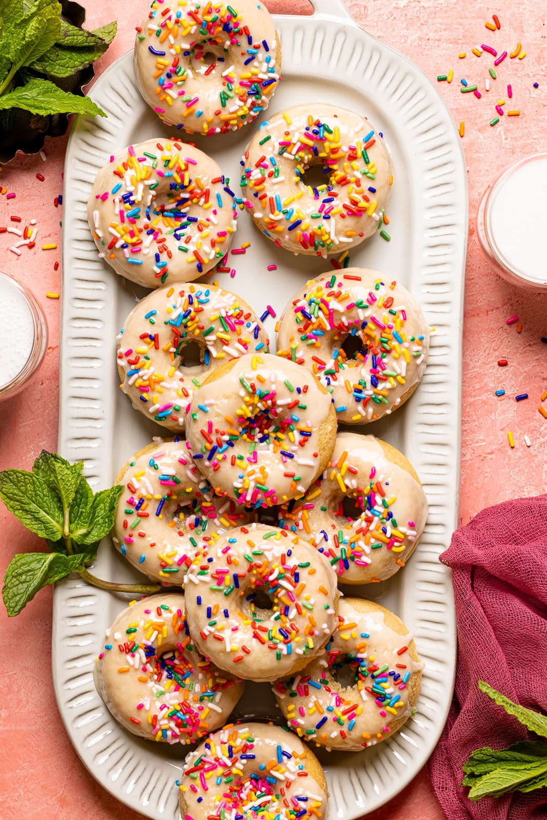 Frosted donuts on a long platter on a pink table with cups of milk, garnish of herbs, and a dark pink napkin.