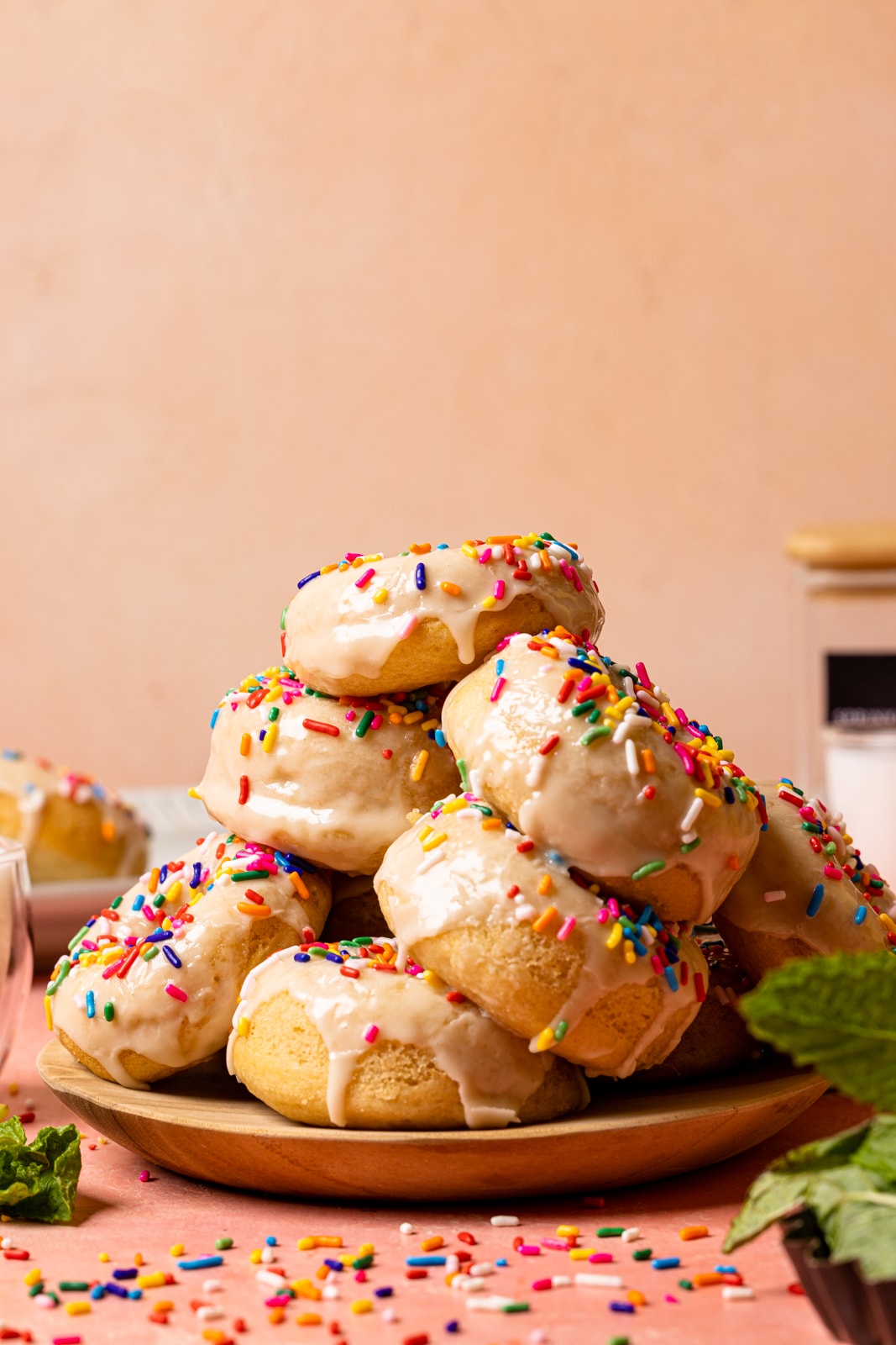 Stacks of frosted donuts on a plate on a pink table with rainbow sprinkles and garnish of herbs.