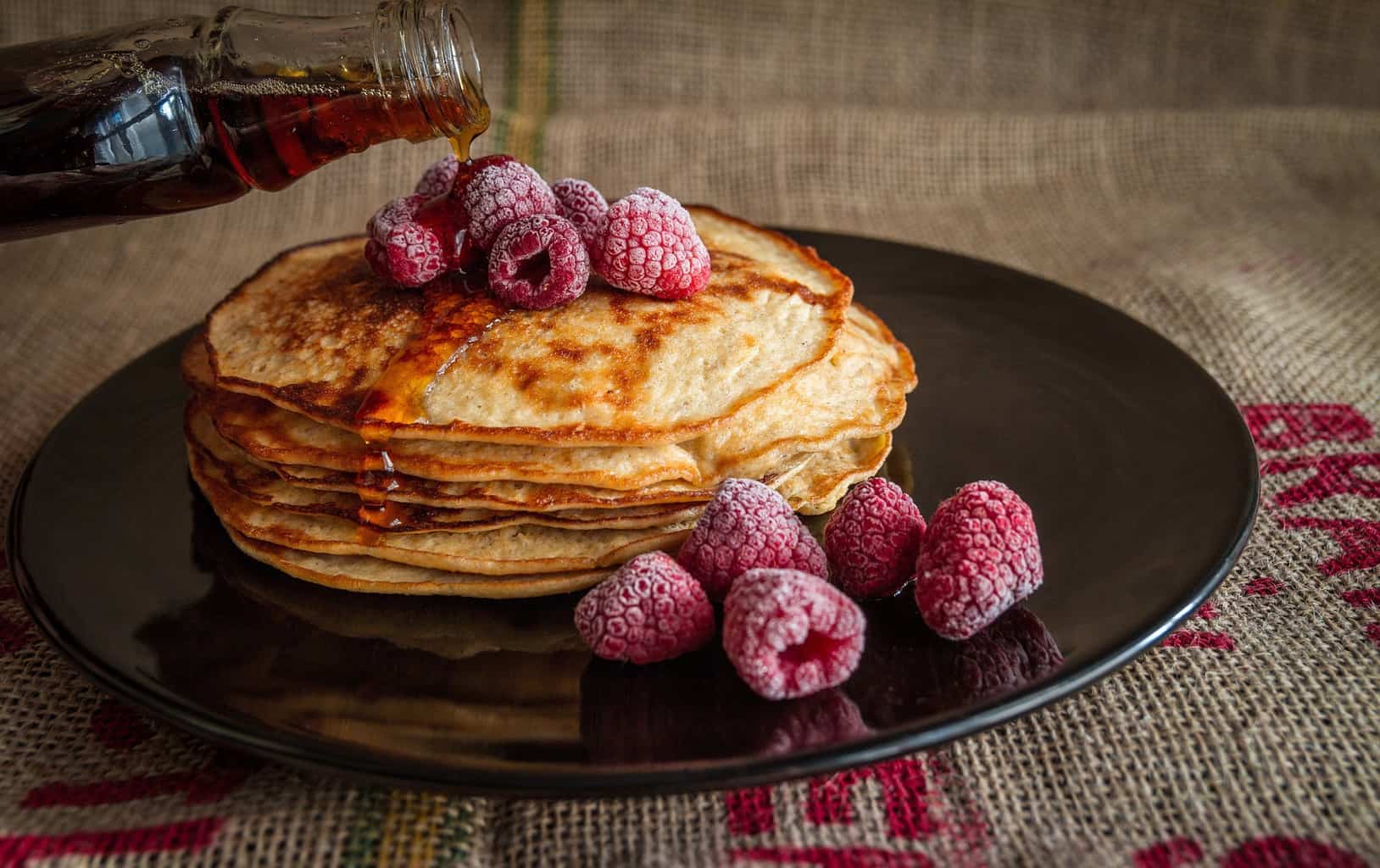 Syrup pouring over a stack of pancakes topped with frozen raspberries.