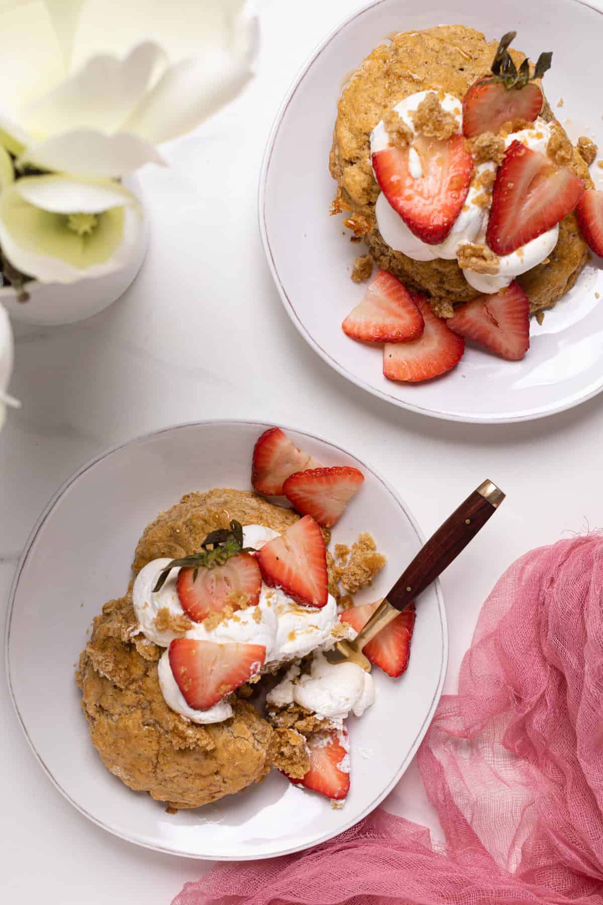 Two servings of Maple Whole Wheat Oatmeal Strawberry Shortcake on white plates with a white flour in the background and pink gauze.