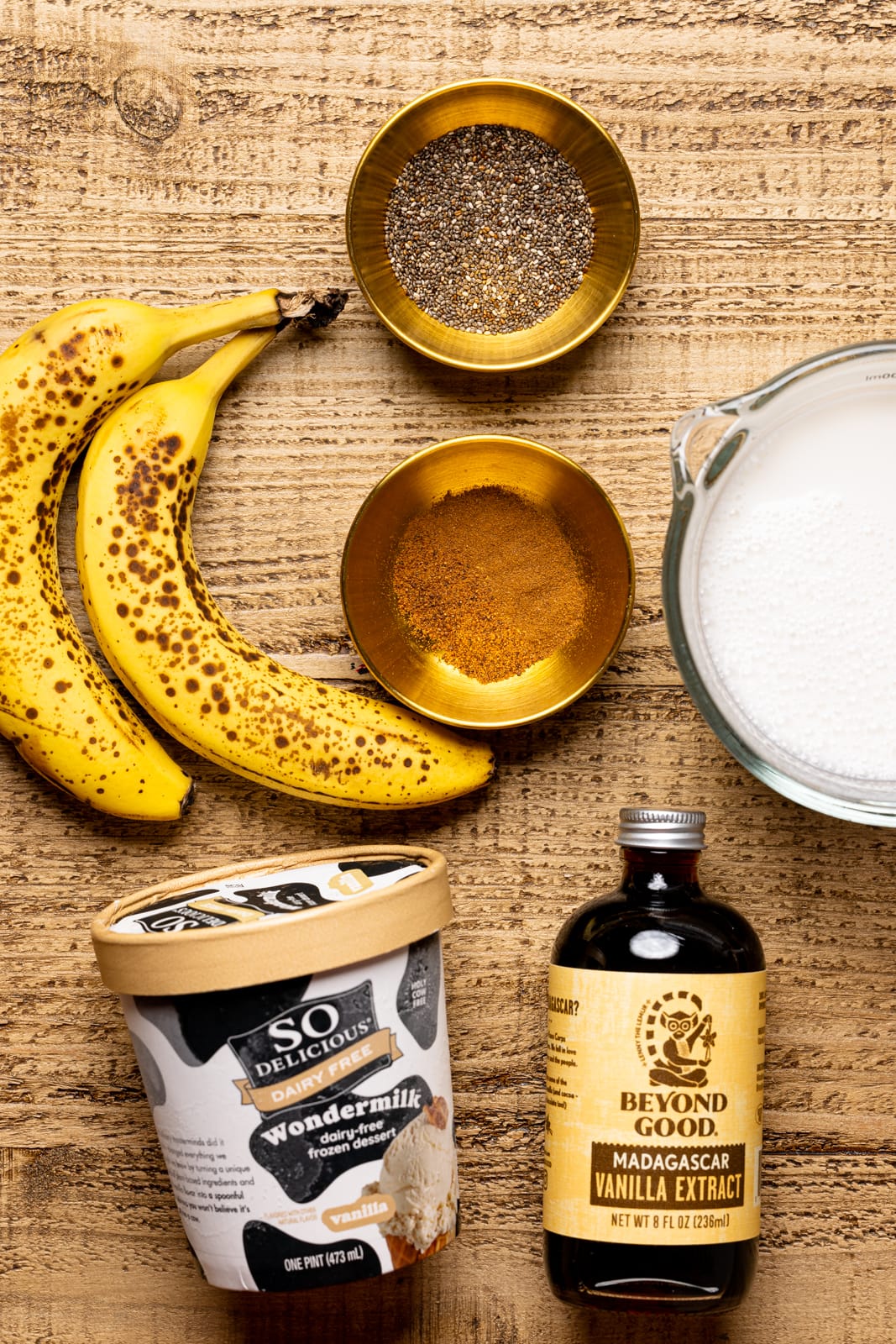 Ingredients on a brown wood table including bananas, vanilla, ice cream, milk, cinnamon, and chia seeds.
