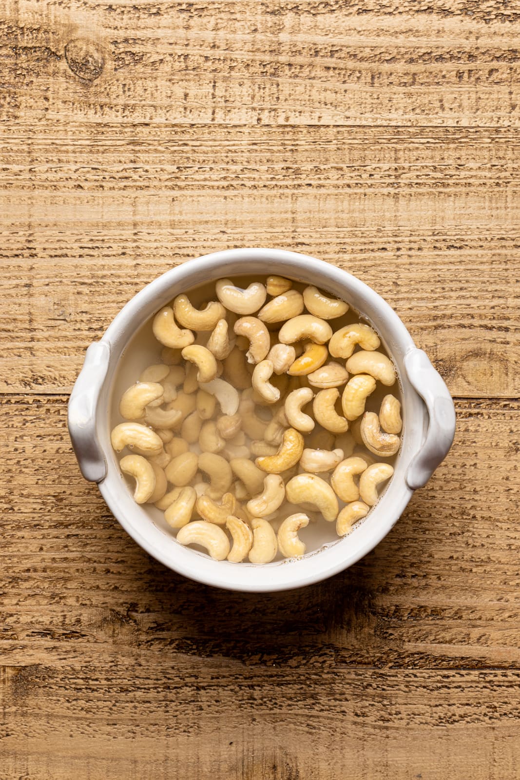 A white bowl with cashews covered with water on a brown table.