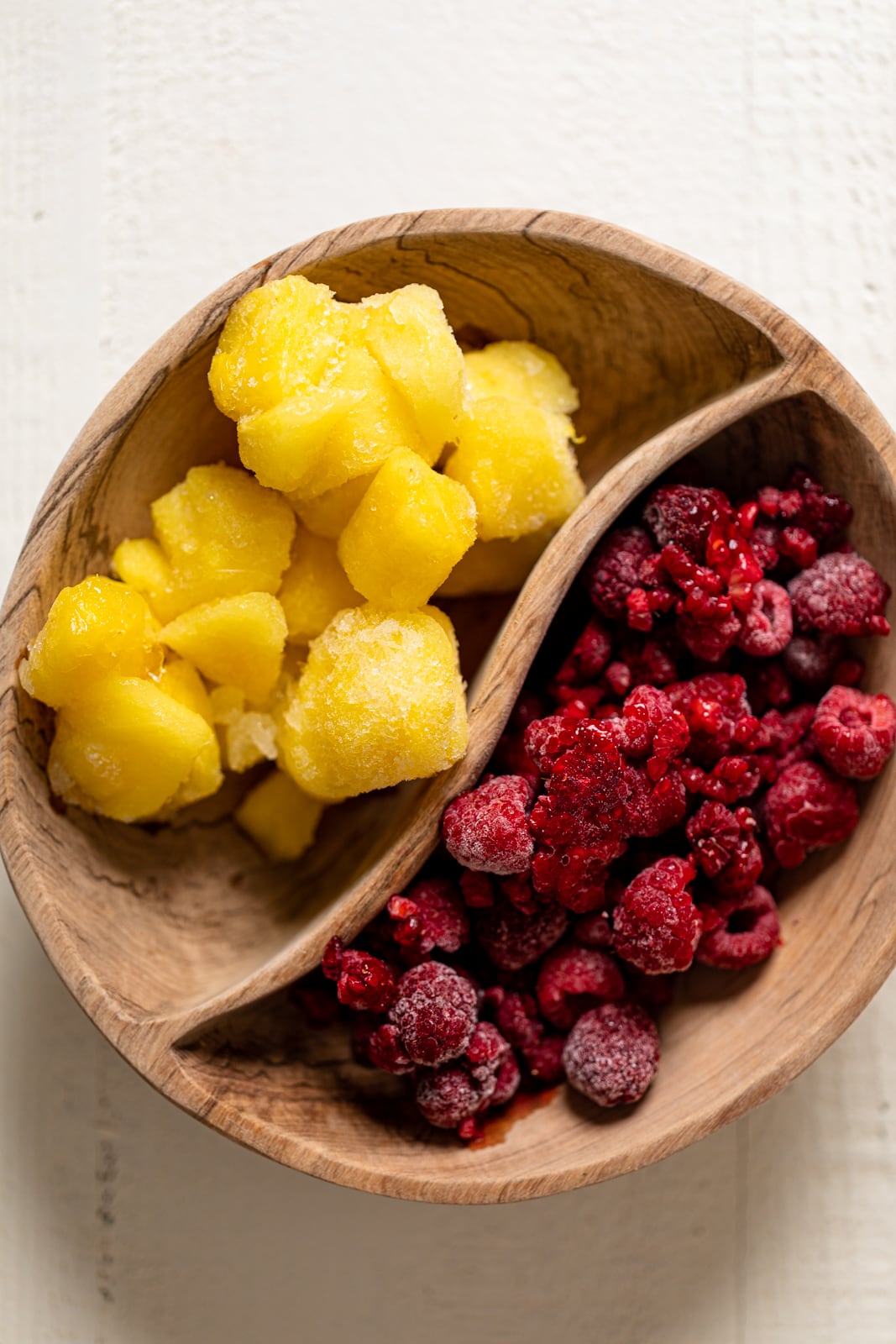 Divided wooden bowl of frozen mango and frozen raspberries.