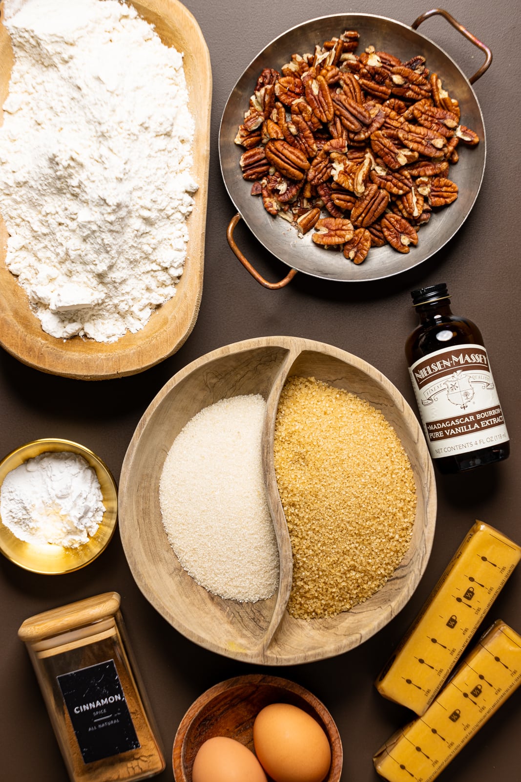 Ingredients on a brown table including flour, sugar, vanilla, eggs, butter, cinnamon, and pecans.