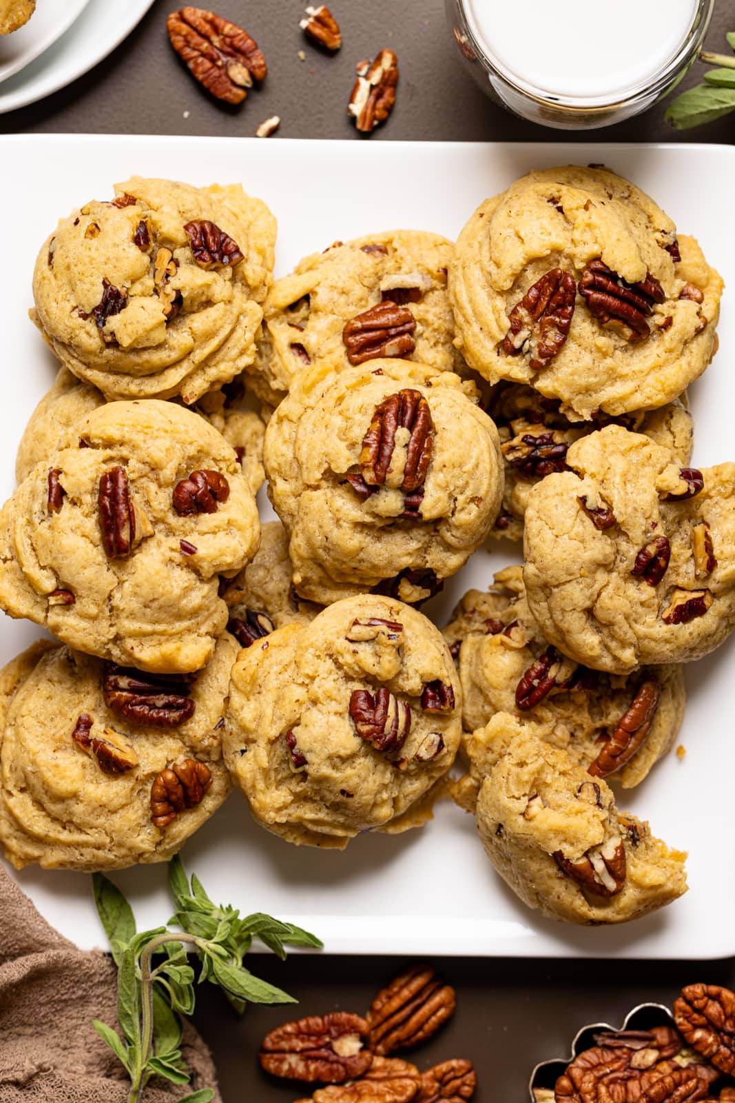 Cookies on a white plate on a brown table with a glass of milk and pecans.