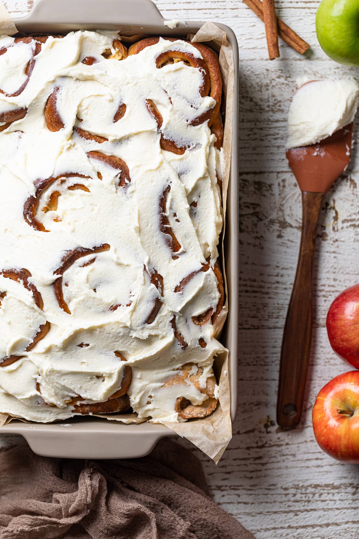 Baking pan of Overnight Vegan Apple Cinnamon Rolls next to apples and a spatula of maple glaze